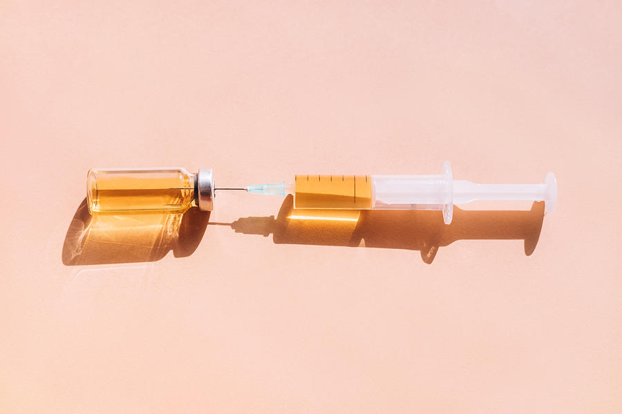 Glass vial of golden vaccine with syringe placed on pastel coral pink background with shadows and light reflections. Flat lay style Photograph by Anna Efetova