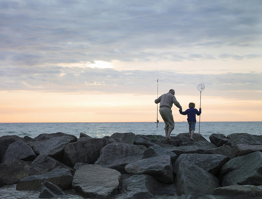 Grandfather and grandson (4-6) walking on rocks with fishing equipment Photograph by Jakob Helbig