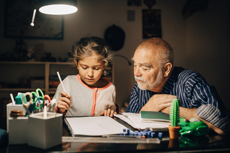 Grandfather looking at granddaughter writing homework on desk in house Photograph by Maskot