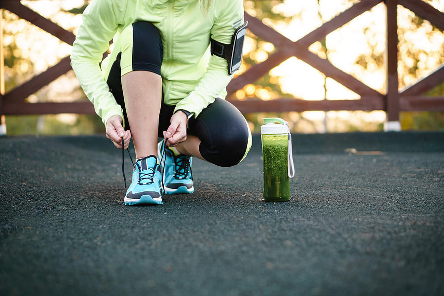 Green detox smoothie cup and woman lacing running shoes before workout. Photograph by Lifemoment