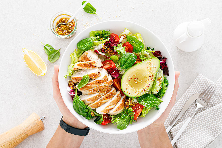 Grilled chicken meat and fresh vegetable salad of tomato, avocado, lettuce and spinach. Healthy and detox food concept. Ketogenic diet. Buddha bowl in hands on white background, top view Photograph by YelenaYemchuk