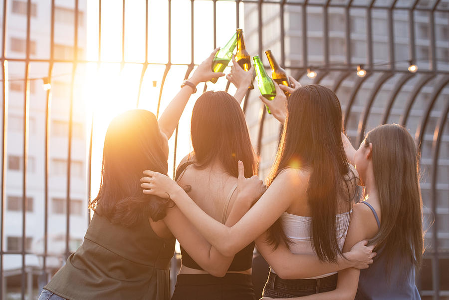 Group of happy beautiful young asian woman friends on back or rear side raised hand hold bottle of beer dance and toast together on outdoor rooftop terrace in evening of outdoors club. Photograph by Mkitina4