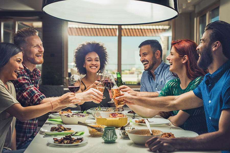 Group of happy friends toasting while eating at dining table. Photograph by Skynesher