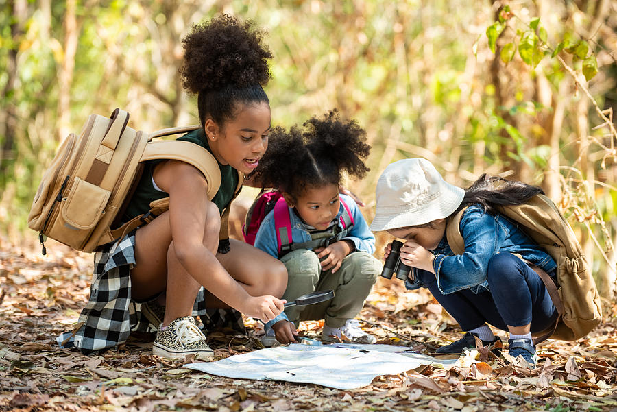 Group of happy pretty little girls hiking together with backpacks and sitting on forest dirt road with looking at the map for exploring the forest. Three kids having fun adventuring in sunny summer day Photograph by CandyRetriever