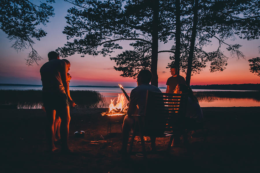 Group of young happy friends sitting by the fire at summer beach, grilling sausages and drinking beer, talking and having fun Photograph by Анна Андреева