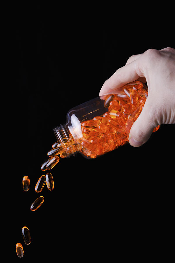 Hand pouring bottle of orange medicine capsules against blue background Photograph by Berkpixels