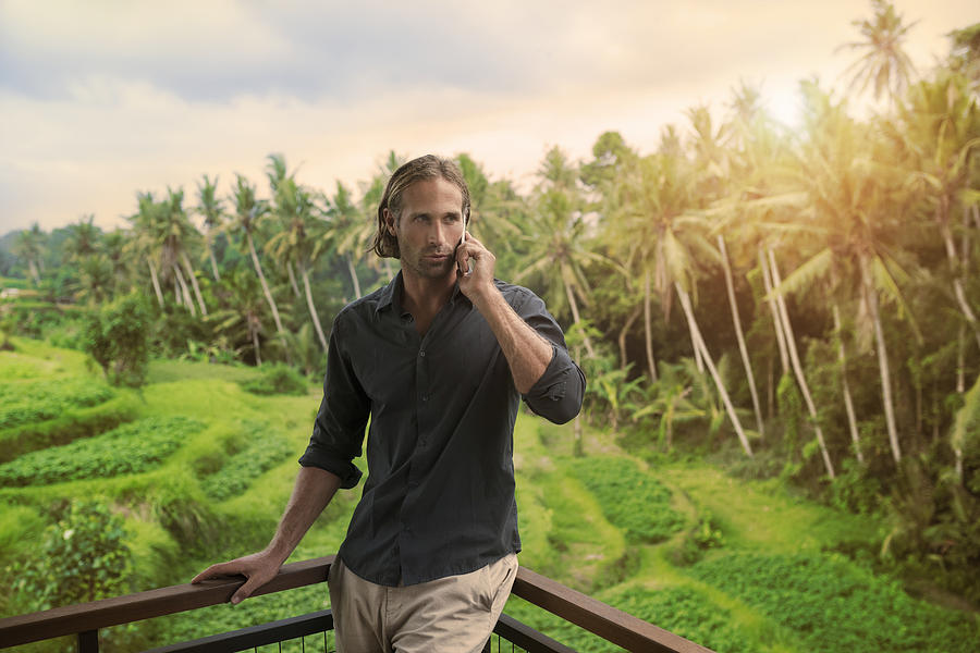 Handsome man leaning on balkony speaking on smartphone with stunning view of tropical landscape Photograph by Westend61