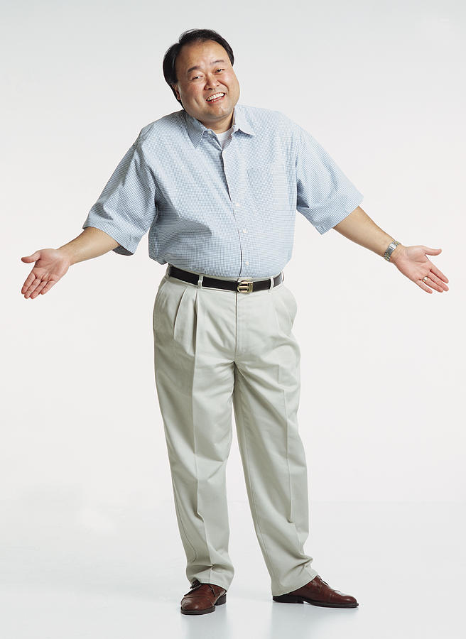 Handsome Middle Aged Asian Adult Male Wearing A Light Blue Short Sleeved Shirt And Cream Colored Slacks Stands With Shrugged Shoulders And Hands In The Air As He Looks At The Camera With A Anxious Smile Photograph by Photodisc