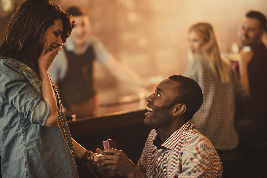 Happy African American man proposing to his girlfriend in a bar. Photograph by Skynesher