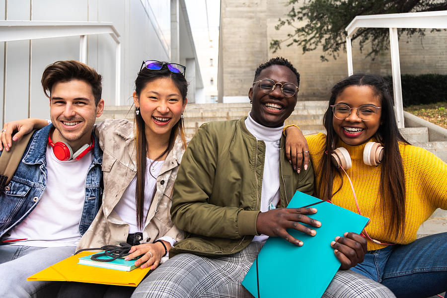 Happy and smiling group of multiracial college student friends looking at camera sitting on stairs outside university building. Photograph by Daniel de la Hoz