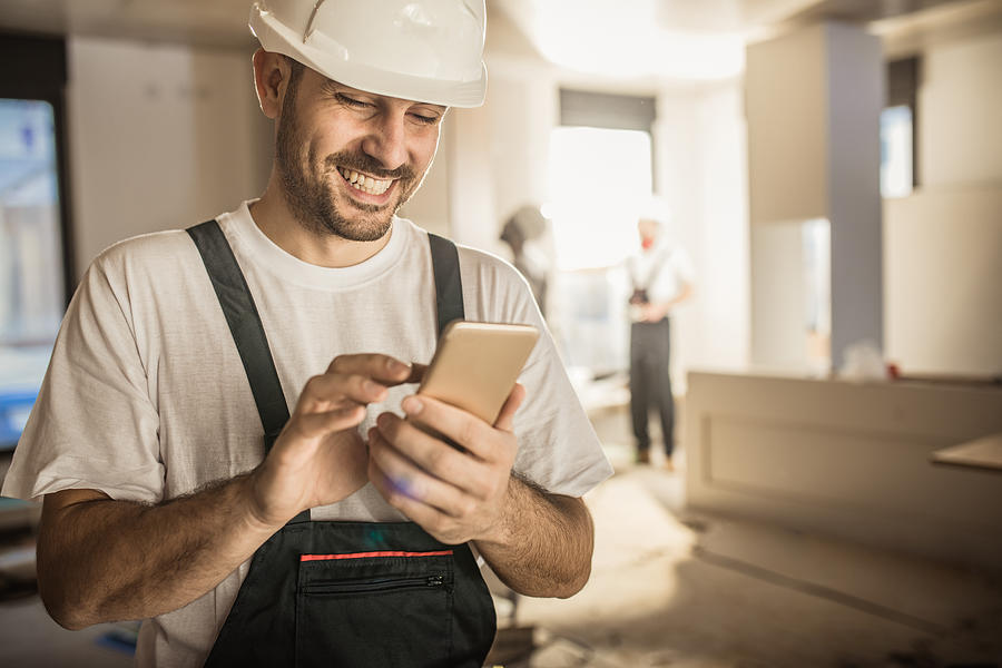 Happy construction worker using cell phone during home renovation. Photograph by Skynesher