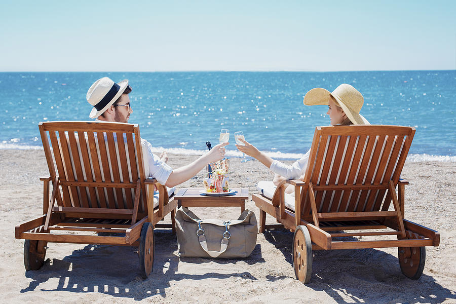 Happy couple clinking their glasses while relaxing on the beach Photograph by Murat Deniz