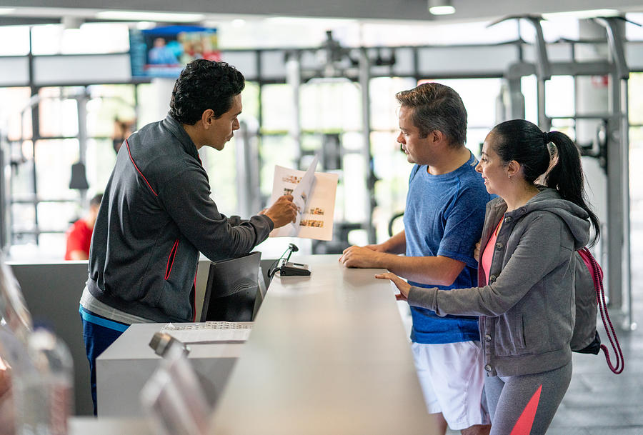 Happy couple registering at the gym while employee explains benefits showing them a catalogue Photograph by Hispanolistic