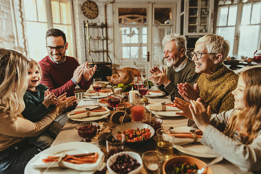 Happy extended family applauding during Thanksgiving meal at dining table. Photograph by Skynesher