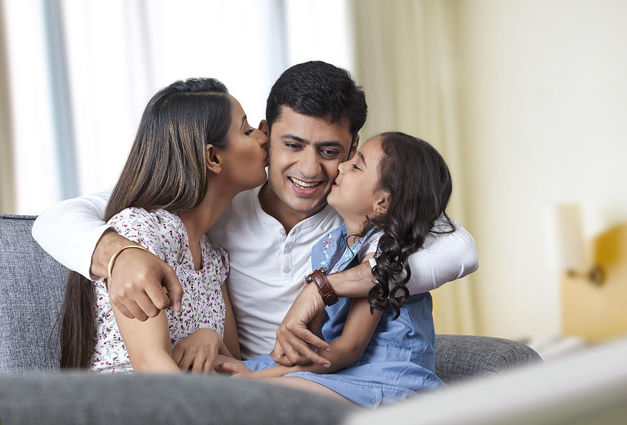 Happy family sitting on sofa mother and daughter kissing father Photograph by IndiaPix/IndiaPicture