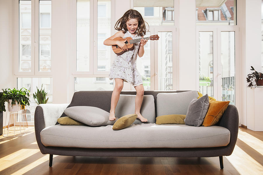 Happy girl playing mini guitar while jumping on a couch in living room at home Photograph by Westend61