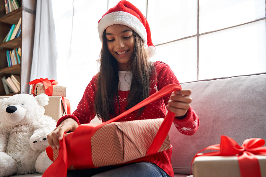 Happy indian kid girl unties red ribbon opens Christmas gift box at home. Smiling cute latin child wearing santa hat receiving xmas New Year present sitting on couch celebrating winter holidays. Photograph by Insta_photos