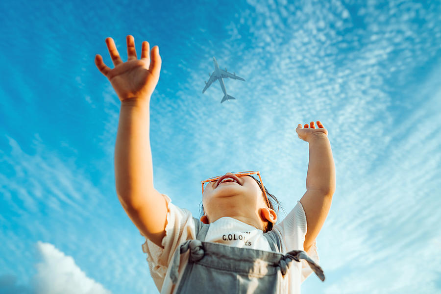 Happy little Asian girl with flower-shaped sunglasses smiling joyfully and raised her hands waving to the aeroplane in the clear blue sky Photograph by D3sign