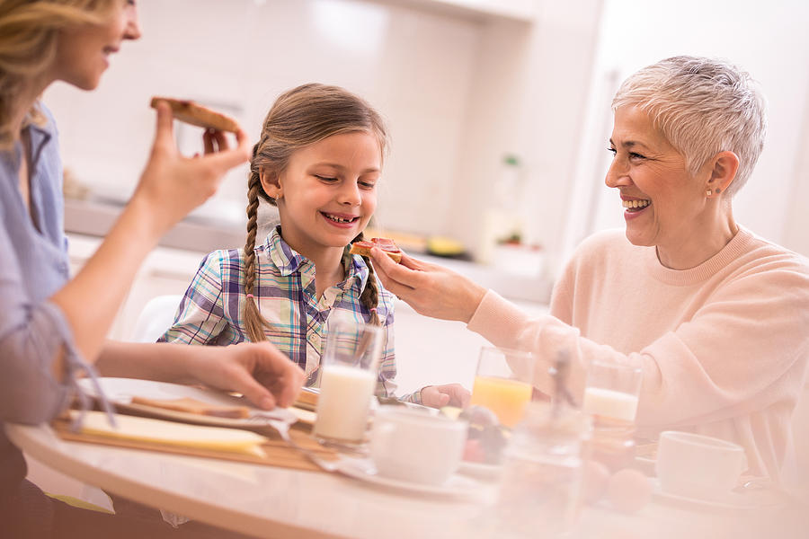 Happy little girl enjoying in breakfast time with her grandmother and mother. Photograph by Skynesher