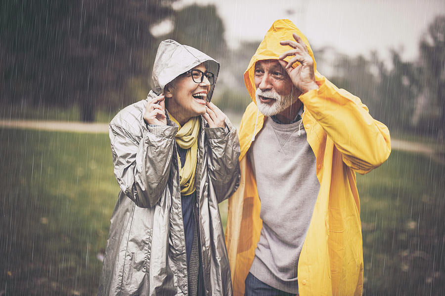 Happy mature couple in raincoats during rain in the park. Photograph by Skynesher