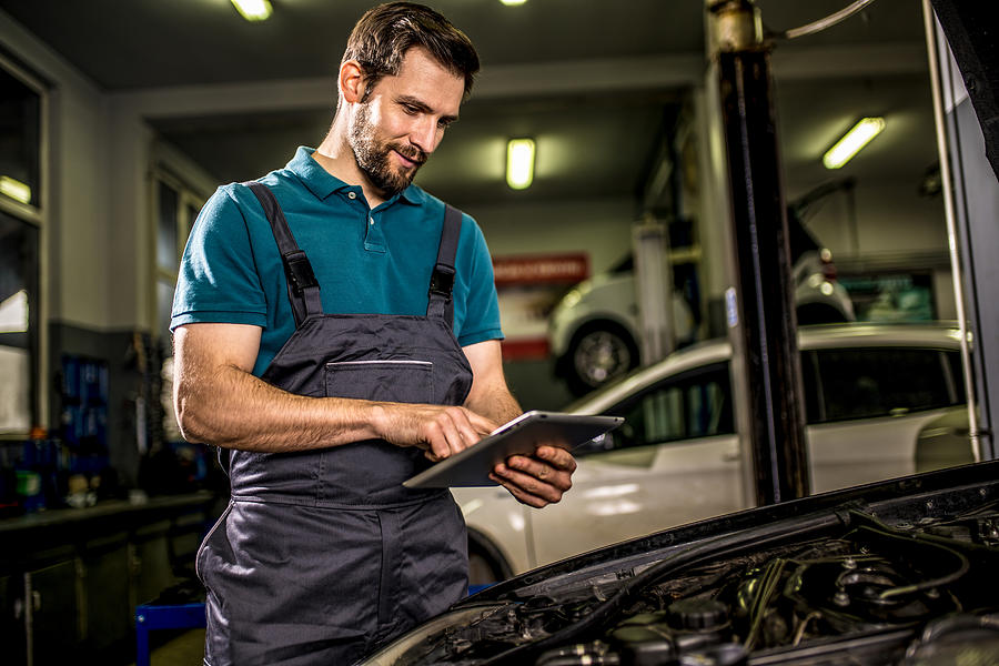 Happy mechanic examining car diagnostics on digital tablet in auto repair shop. Photograph by DjelicS