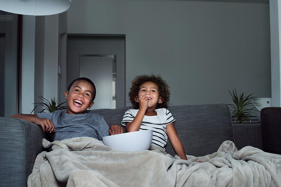 Happy siblings having popcorn while watching TV Photograph by Portra