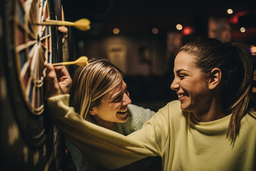 Happy women talking while removing darts from dartboard. Photograph by Skynesher