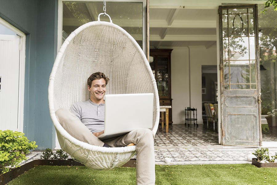 Happy young man using laptop on swing chair Photograph by Portra