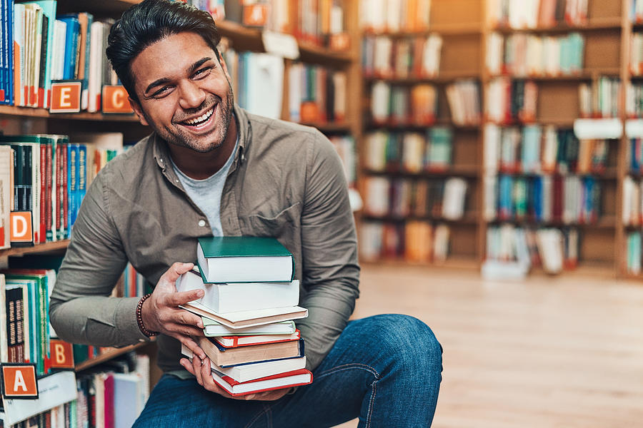 Happy young man with a pile of books in a bookstore Photograph by Pixelfit