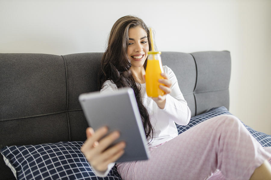 Happy young woman working from home and drinking juice in bed Photograph by DjordjeDjurdjevic