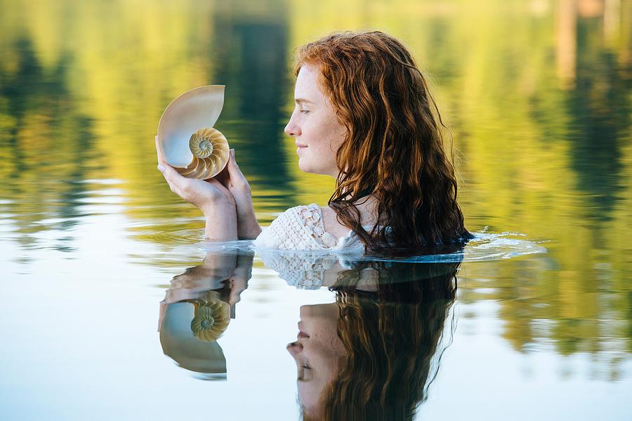 Head and shoulders of young woman with long red hair in lake gazing at seashell Photograph by Pete Saloutos