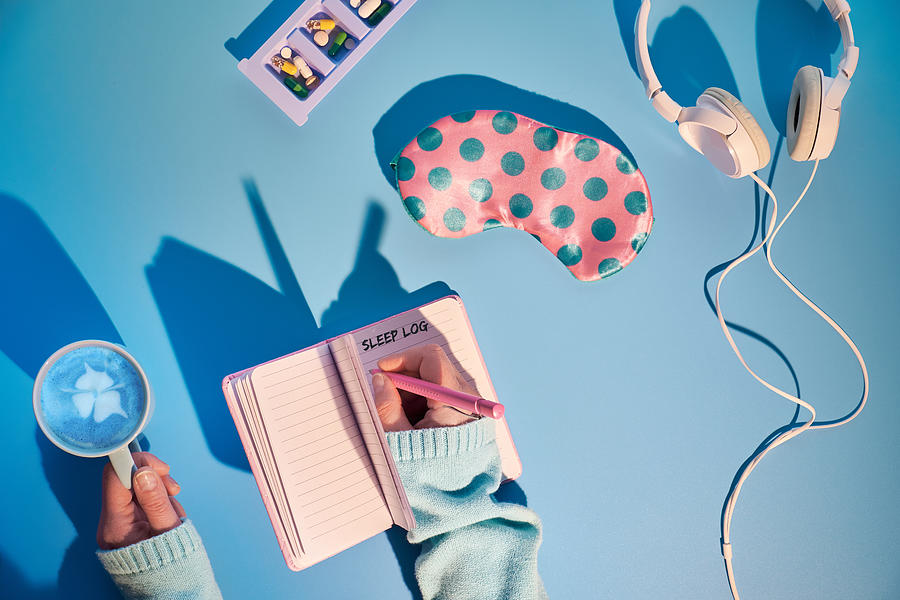 Healthy night sleep creative concept in pink and blue. Sleep mask, pink with green polka dots, white earphones, calming pills against insomnia, sleep log notebook. Blue mint background, long shadows. Photograph by Anyaivanova