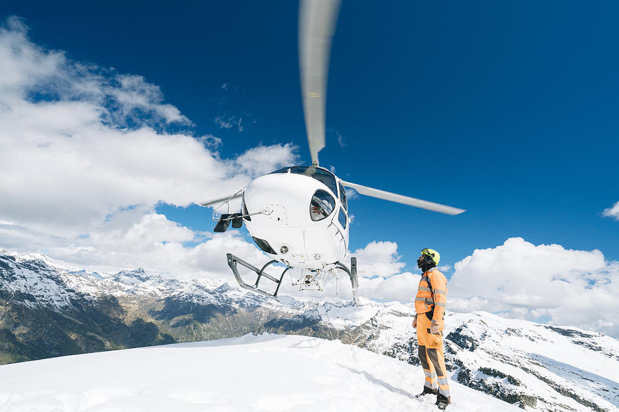 Helicopter lands on mountain summit, ground crew member guides below Photograph by Ascent/PKS Media Inc.