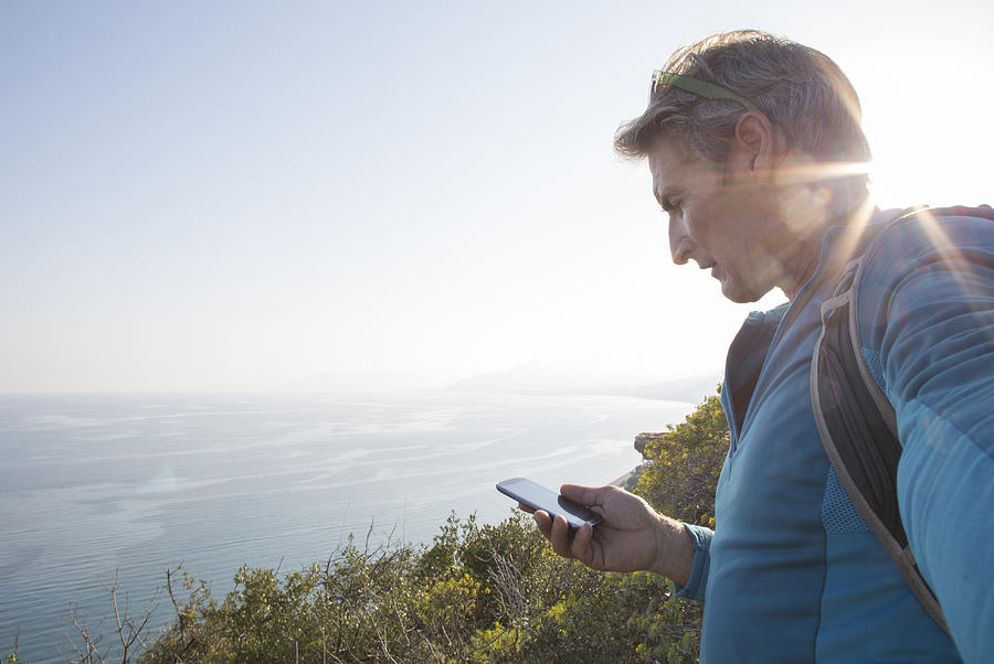 Hiker checks smart phone/GPS on hillside, lake Photograph by Ascent/PKS Media Inc.