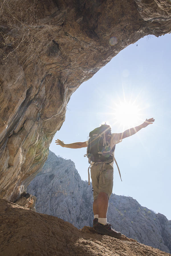 Hiker pauses at cave opening, arms outstretched Photograph by Ascent/PKS Media Inc.