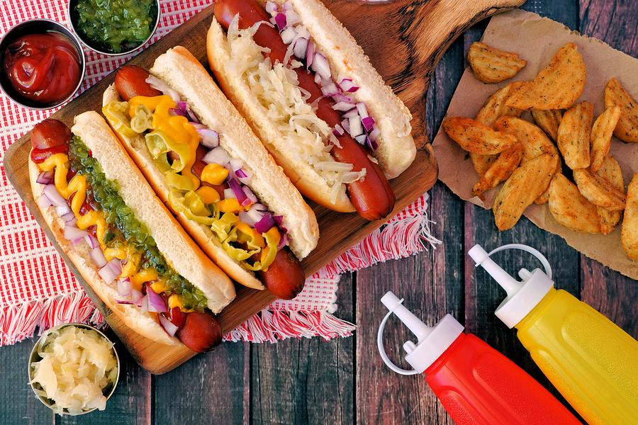 Hot dogs with assorted toppings and potato wedges, overhead scene on wood Photograph by Jenifoto