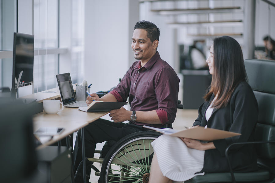 Indian white collar male worker in wheelchair having cheerful discussion conversation with his female asain chinese colleague coworking in creative office workstation beside window Photograph by  Edwin Tan 