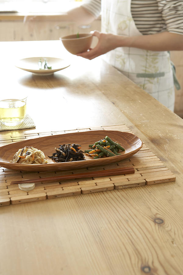 Japanese style vegetables in wooden tray with chopsticks on wooden table, woman preparing food in background Photograph by Ultra F