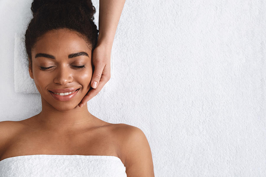 Joyful african girl having face massage at spa Photograph by Prostock-Studio