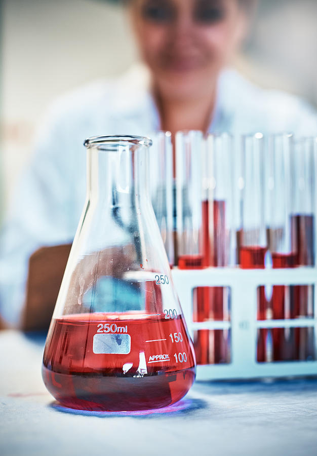 Lab flask and test tubes with red liquid, scientist behind Photograph by RapidEye