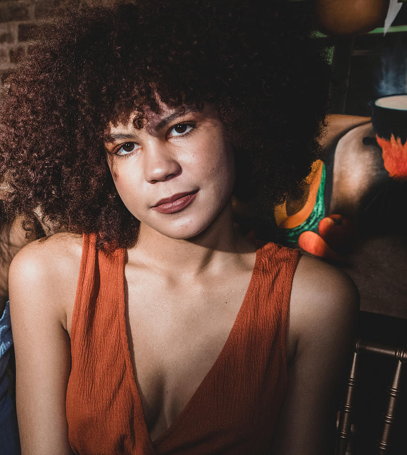 Latina teen sitting at dinner table looking at the camera. Teen has an auburn afro and is wearing an orange colored top. Brick wall and painting in the background. Photograph by Evelyn Martinez