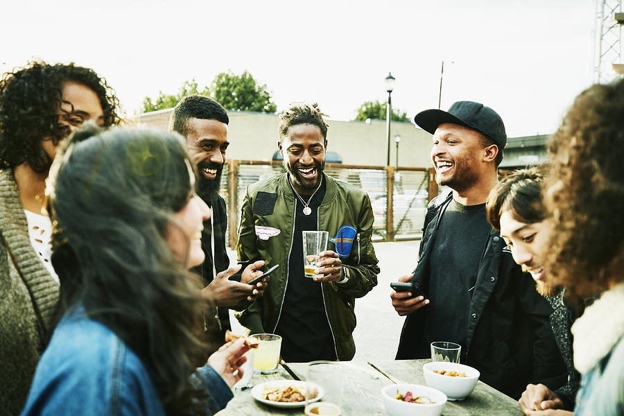 Laughing friends sharing drinks and food at outdoor bar Photograph by Thomas Barwick