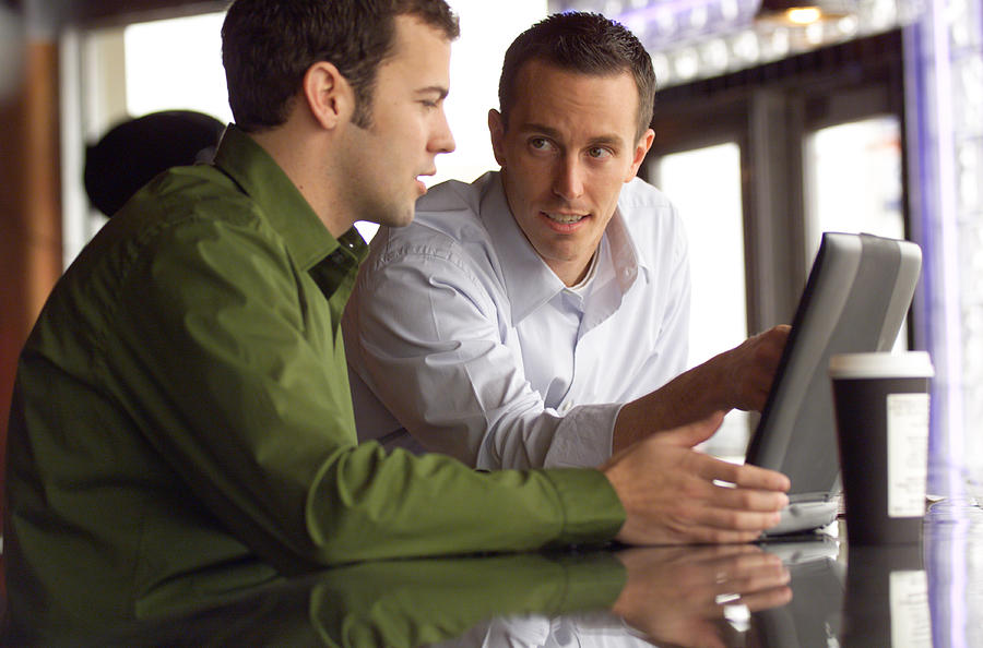 Lifestyle Photo Of Two Caucasian Male Friends As They Sit And Talk Around A Laptop Computer Photograph by Photodisc