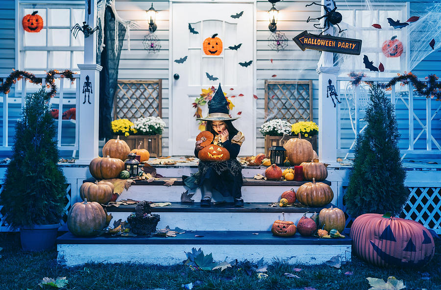 Little girl in witch costume sitting on the stairs in front of the house and holding Jack-o-Lantern Pumpkins on Halloween trick or treat Photograph by Valentinrussanov