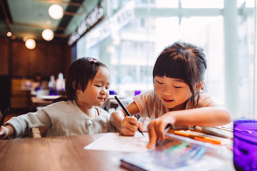 Little girl teaching her little toddler sister to draw joyfully in a restaurant. Photograph by Images By Tang Ming Tung