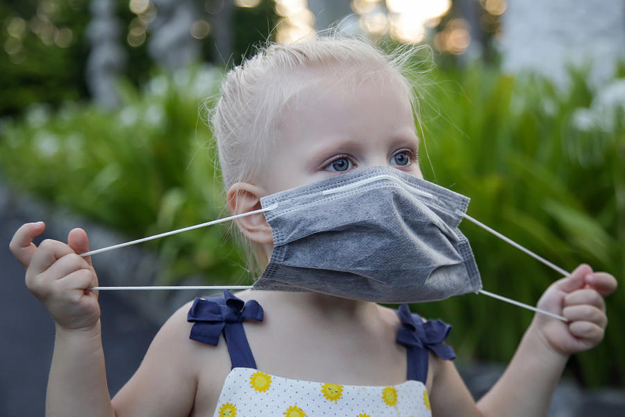 Little toddler girl trying to put medical protective mask. Candid outdoor portrait of child with medical mask. Corona virus outbreak or air pollution concept. Photograph by Triocean