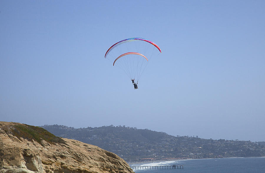 Looking up at two paragliders; cliff, ocean and pier in foreground; coastline and blue sky beyond Photograph by Timothy Hearsum