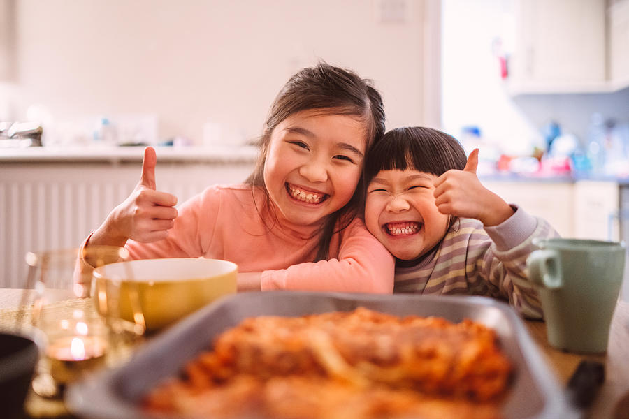 Lovely little sister giving thumbs-up & smiling joyfully at the camera while having meal at home Photograph by Images By Tang Ming Tung