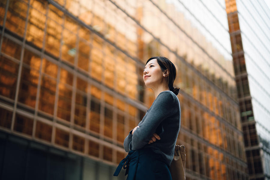 Low angle portrait of confident and successful young Asian businesswoman standing against contemporary corporate skyscrapers in financial district. Female leadership and determined to success Photograph by D3sign