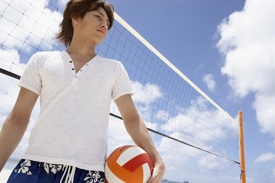 Low Angle View of a Young Man Standing by a Volleyball Net on the Beach and Holding a Ball Photograph by Dex